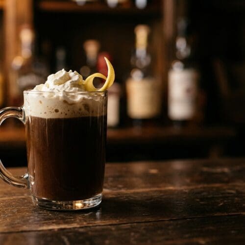 Buttsex cocktail in a clear glass coffee mug topped with whipped cream and a lemon twist on a dark wooden bar